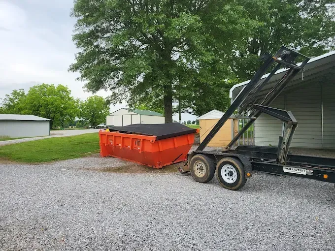 Orange dumpster beside a black trailer hitch on a gravel driveway, with trees and a house in the background