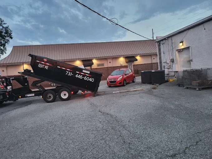 Red utility truck dumping debris in an industrial lot beside a red car and warehouse building at dusk