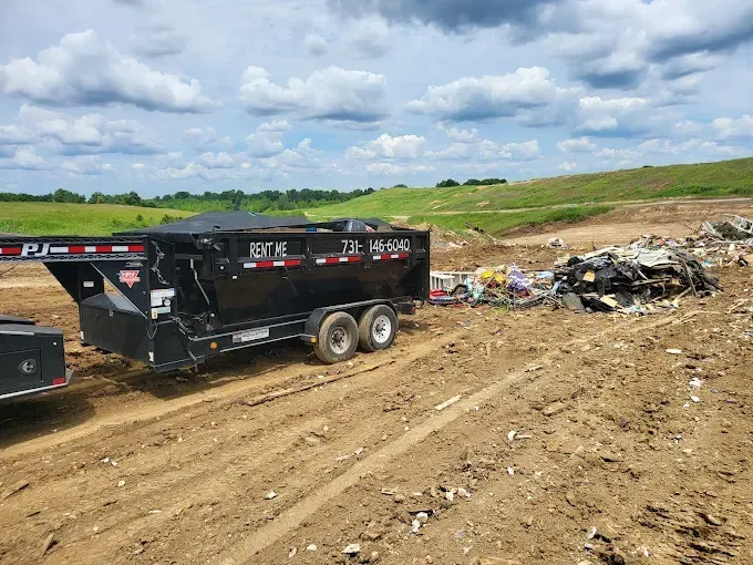 Black dump trailer on muddy dirt road beside piles of debris under a cloudy sky.