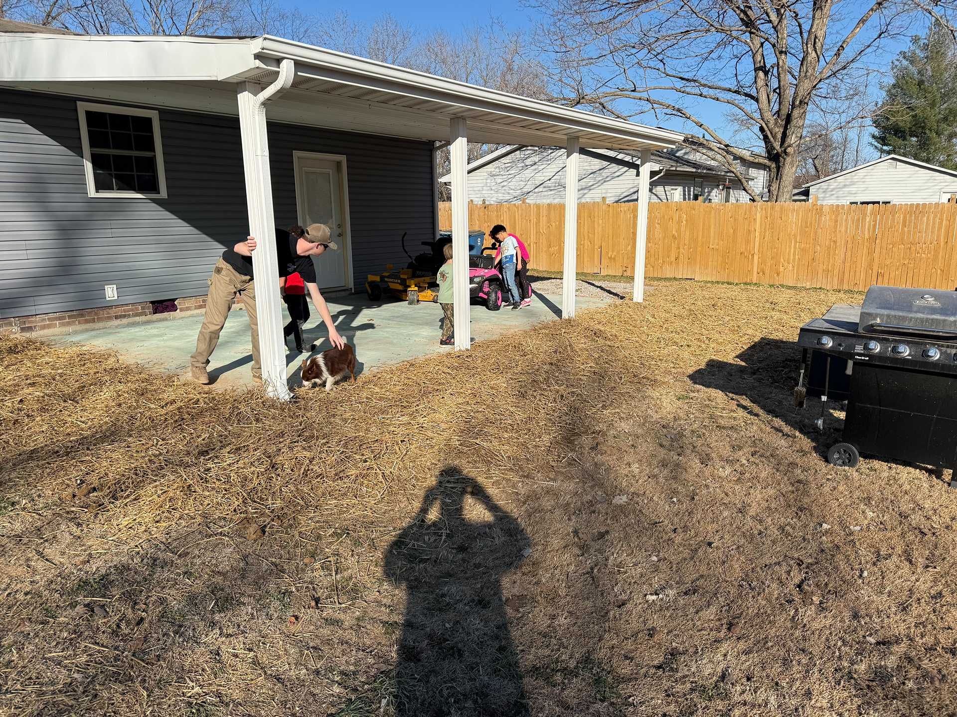 Children playing under a white backyard patio beside a gray house, with leaf-covered ground and a fence.