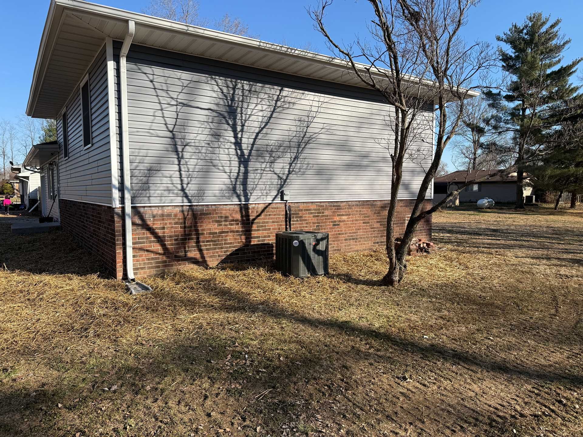 Single-story house with white siding and brick foundation, leafless trees, and an outdoor AC unit in a yard.