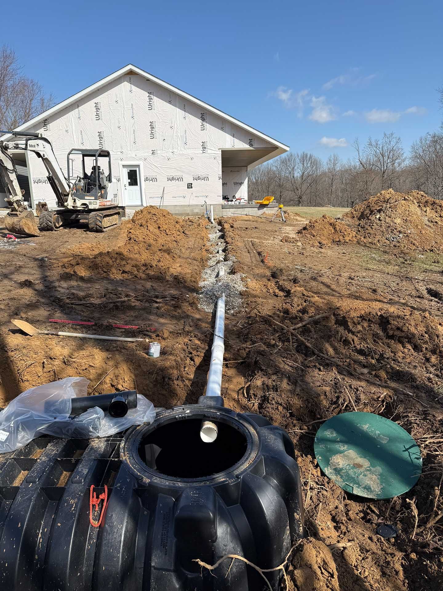 Construction site with excavator, dug trench, and black septic tank beside a white house.