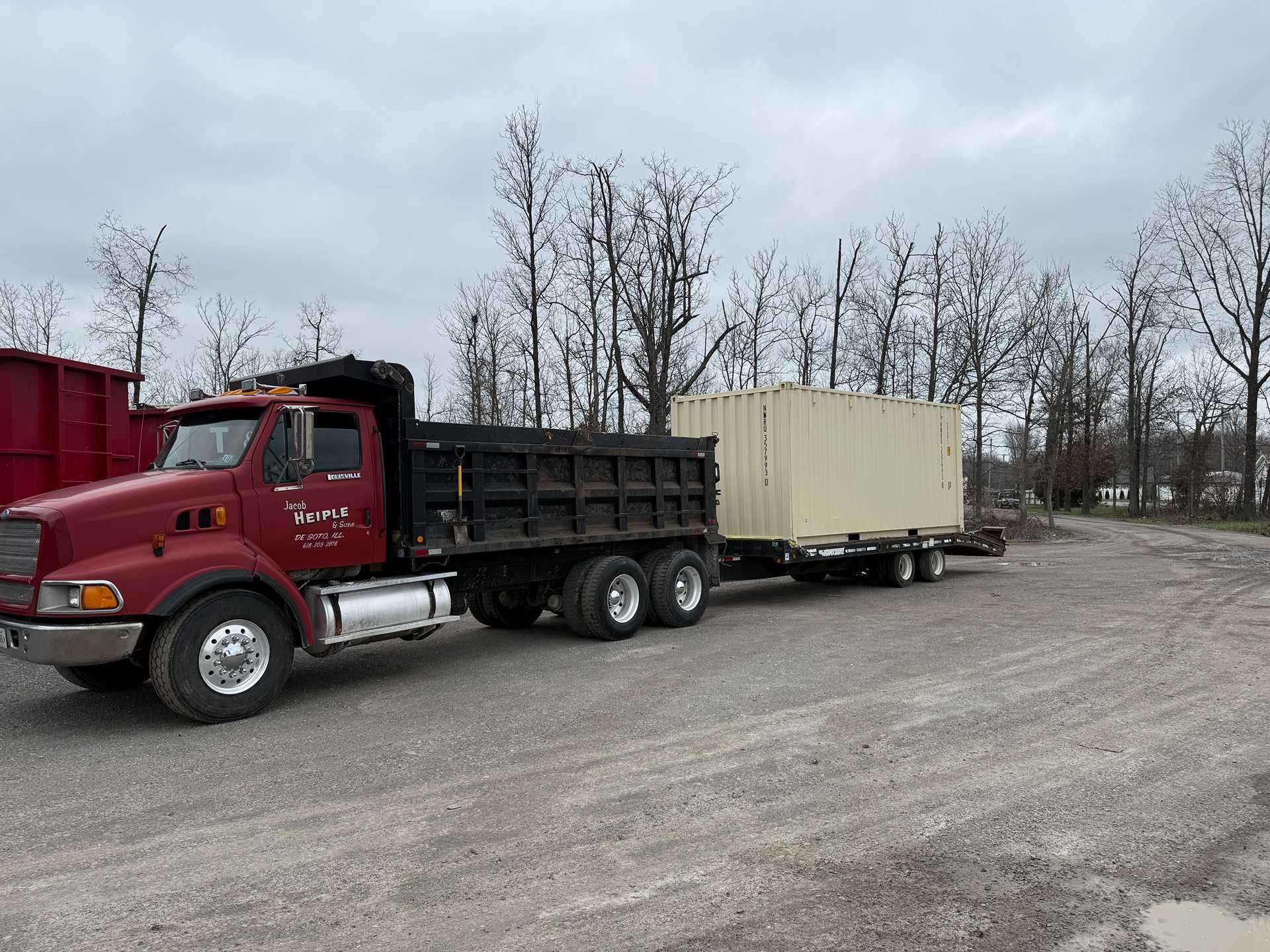 Red dump truck hauling a beige trailer on a gravel lot beside leafless trees.