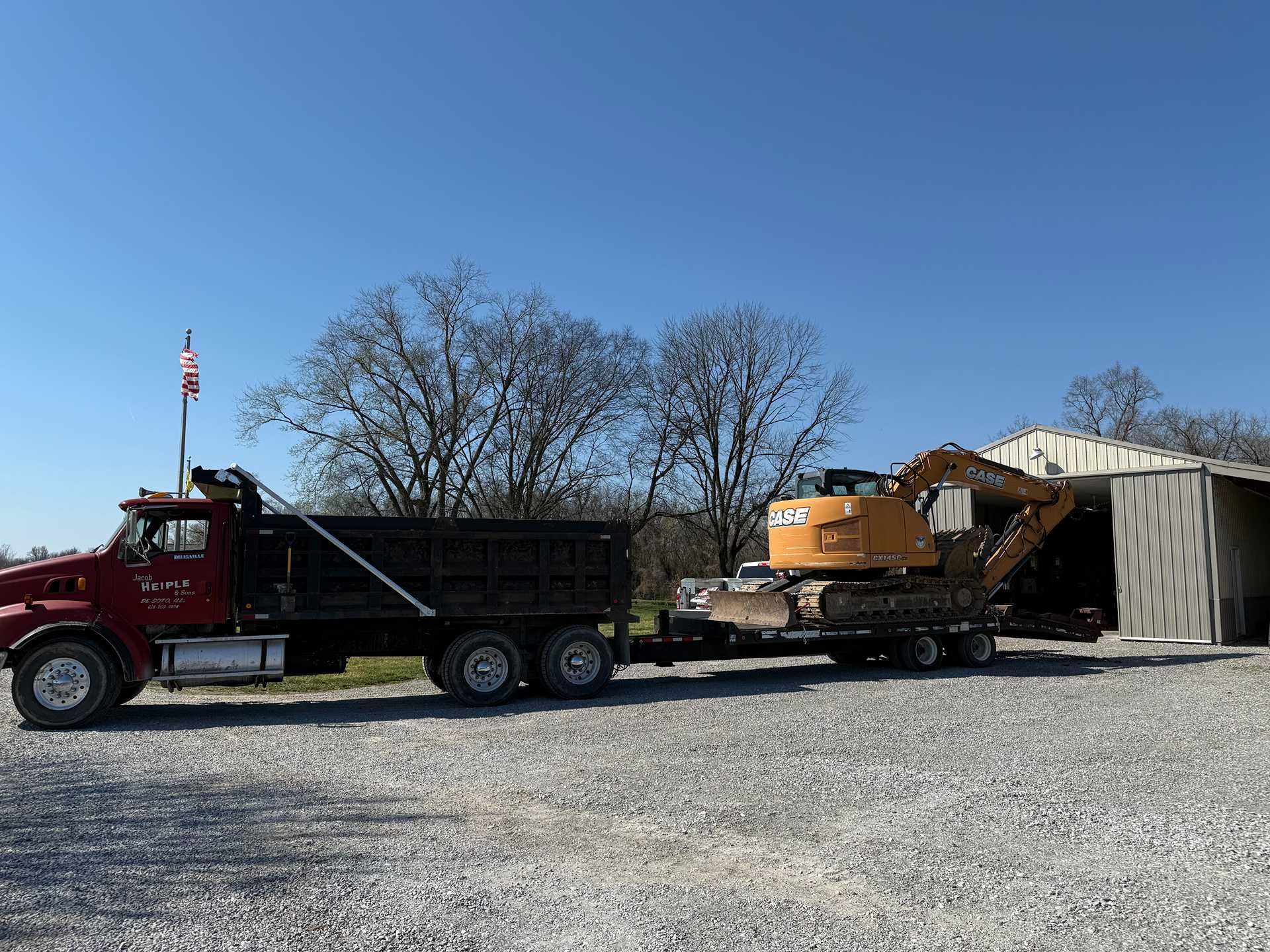 Red dump truck hauling a yellow excavator beside a metal shed on a gravel lot
