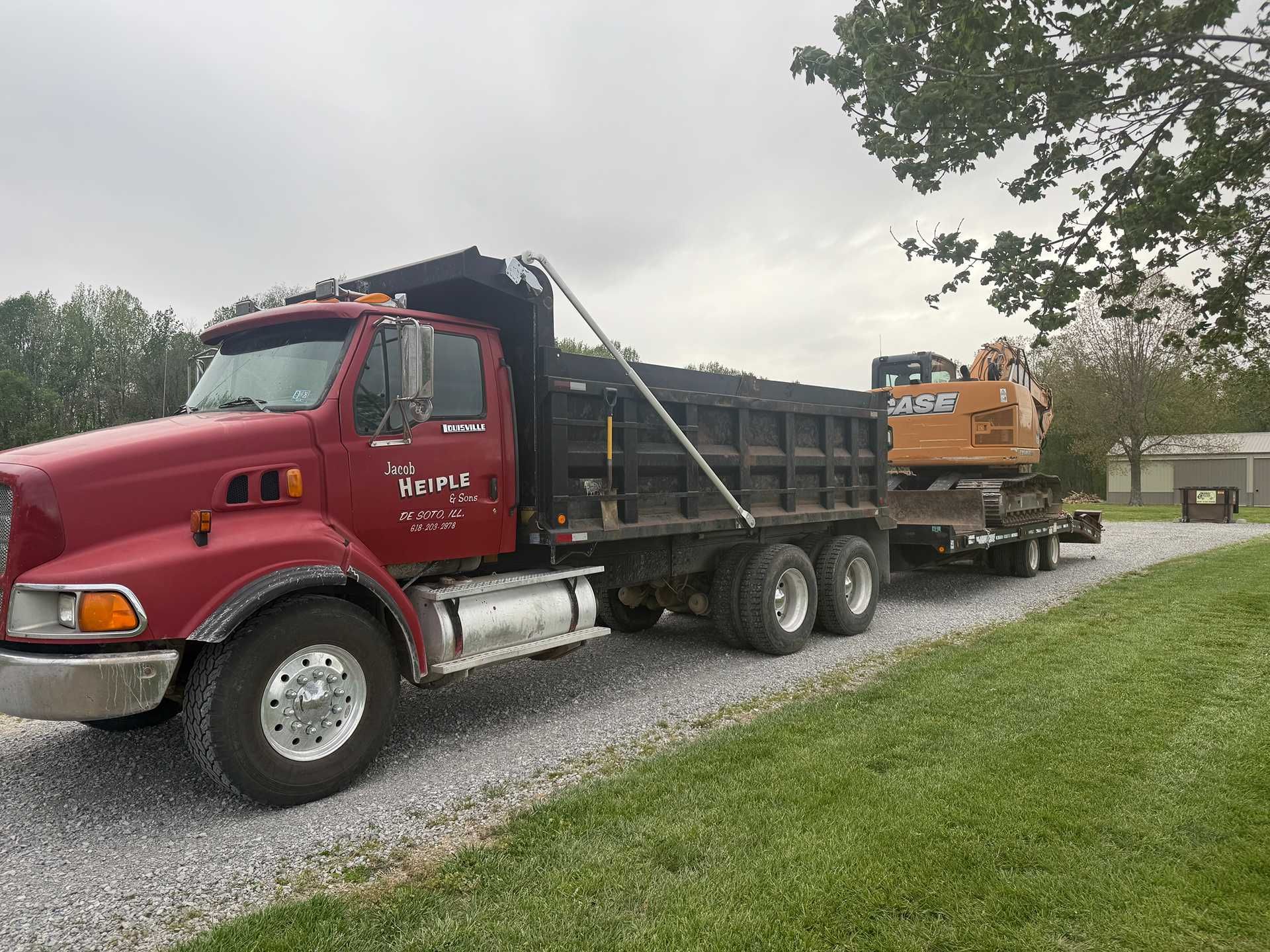 Red dump truck hauling an orange excavator on a trailer beside a grassy roadside.