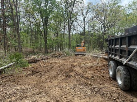Excavator leveling a dirt lot beside a black dump trailer in a wooded area