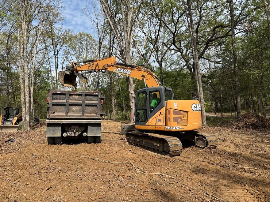 Orange excavator loading dirt into a dump truck in a wooded clearing