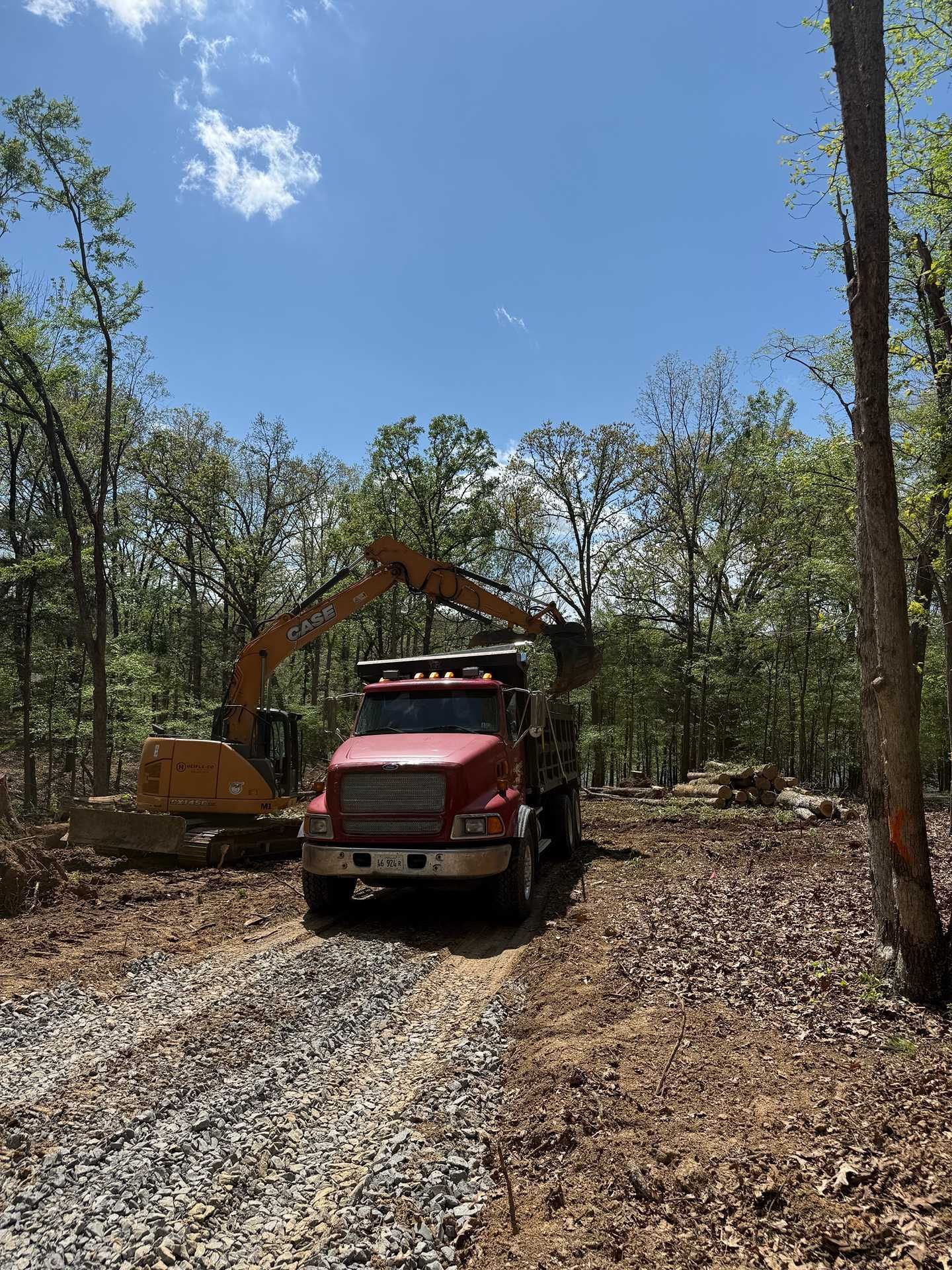 Red dump truck and excavator on a gravel path in a wooded area under a blue sky