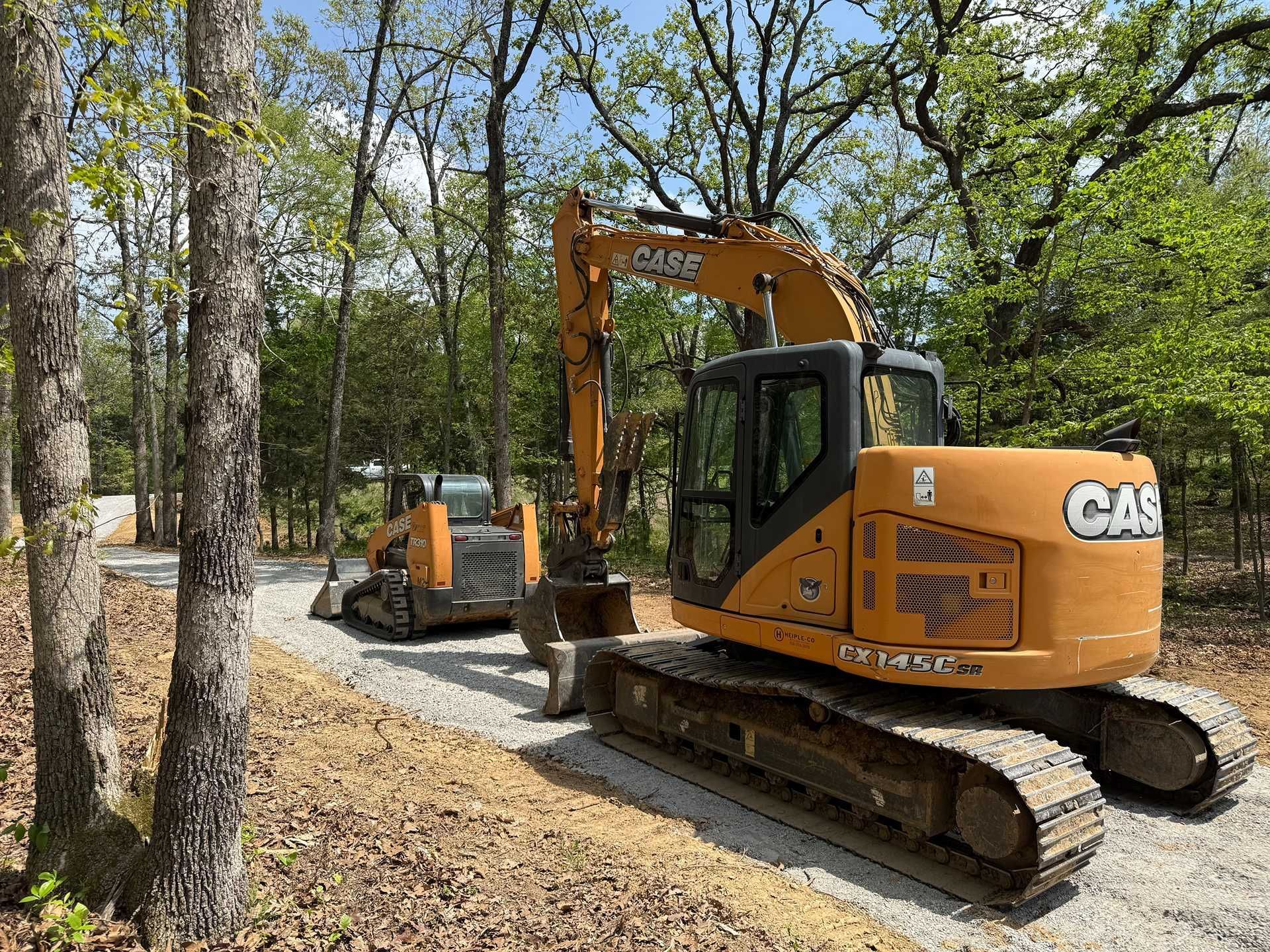 Two yellow excavators on a wooded dirt road in spring greenery
