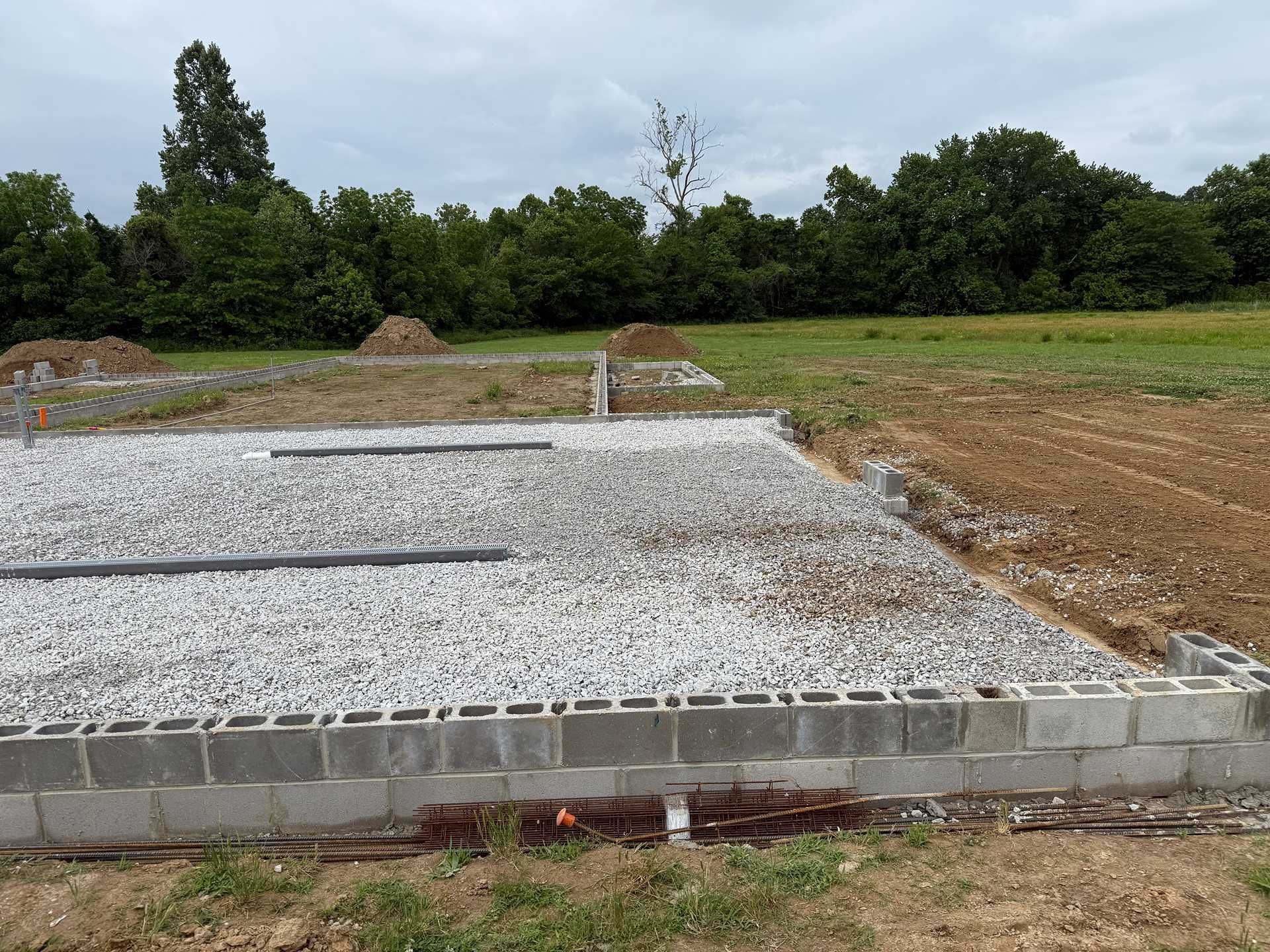 Gravel-covered foundation slabs at a construction site with trees and open field in the background