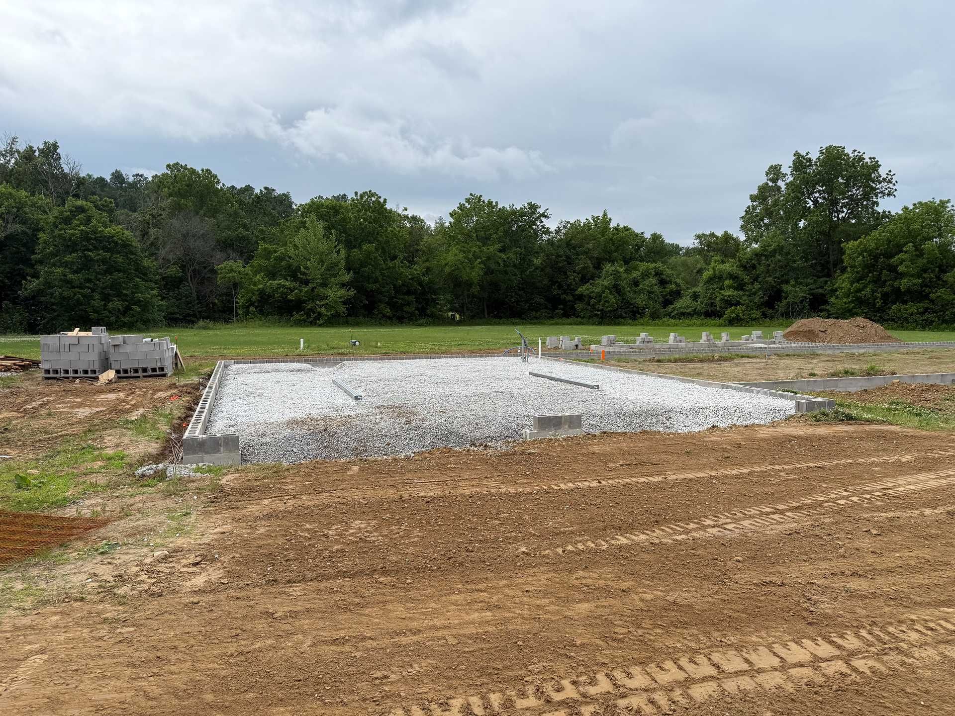 Gravel foundation and dirt lot at a construction site under cloudy sky, with trees in the background