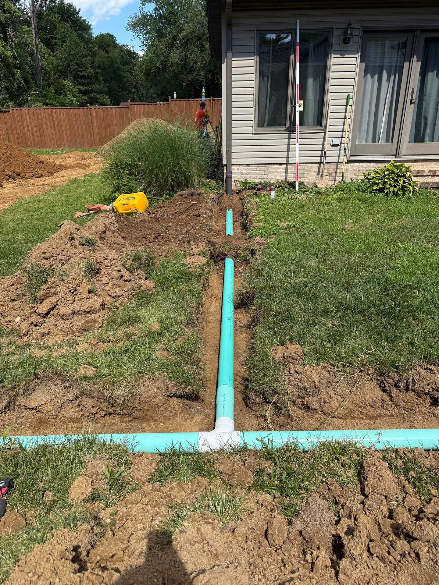 Trench with blue pipe leading to house foundation in a grassy yard, with excavated dirt and a yellow tool nearby