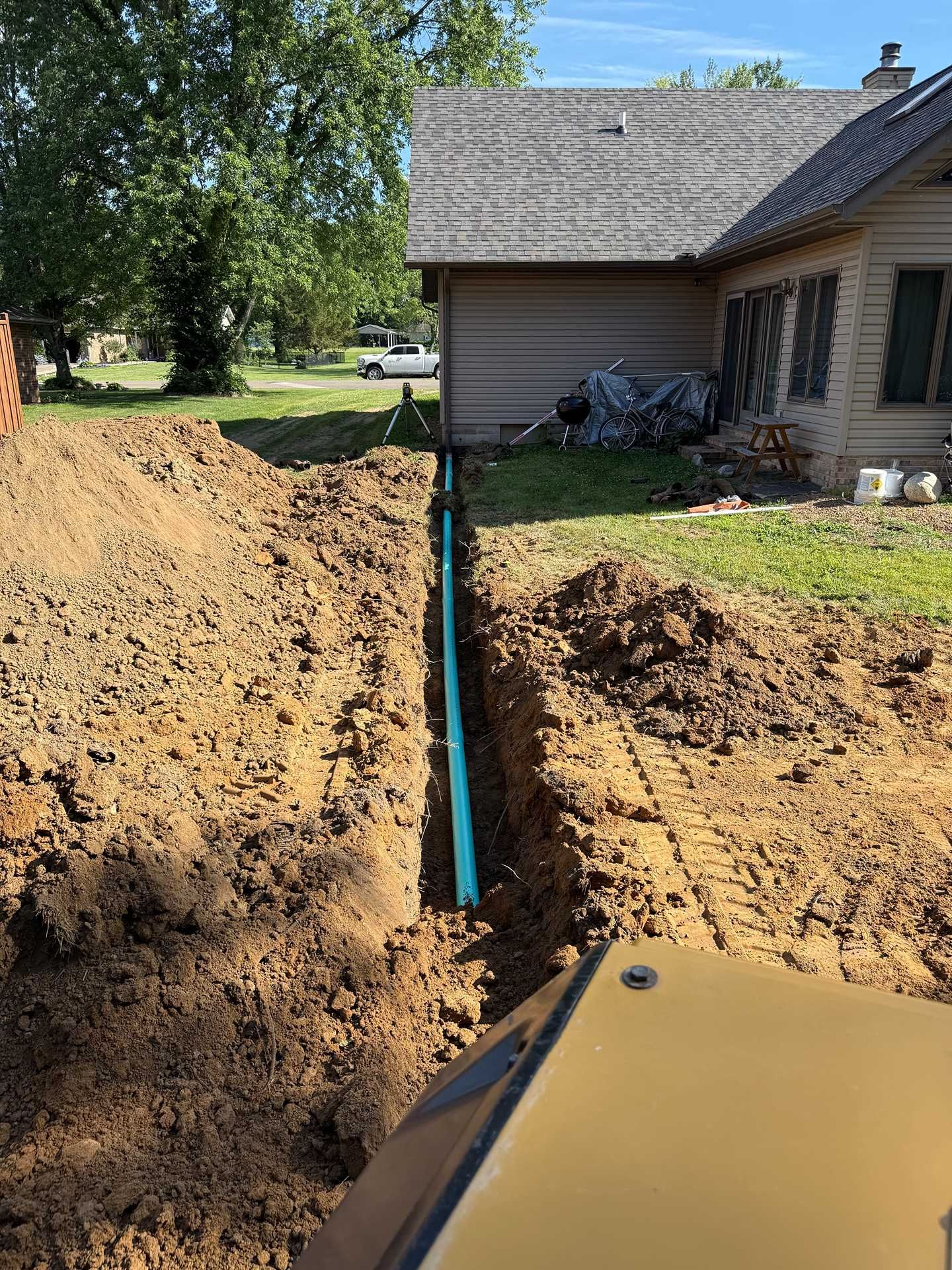 Trench beside a house with a blue pipe, dirt piles, and muddy water in the foreground.