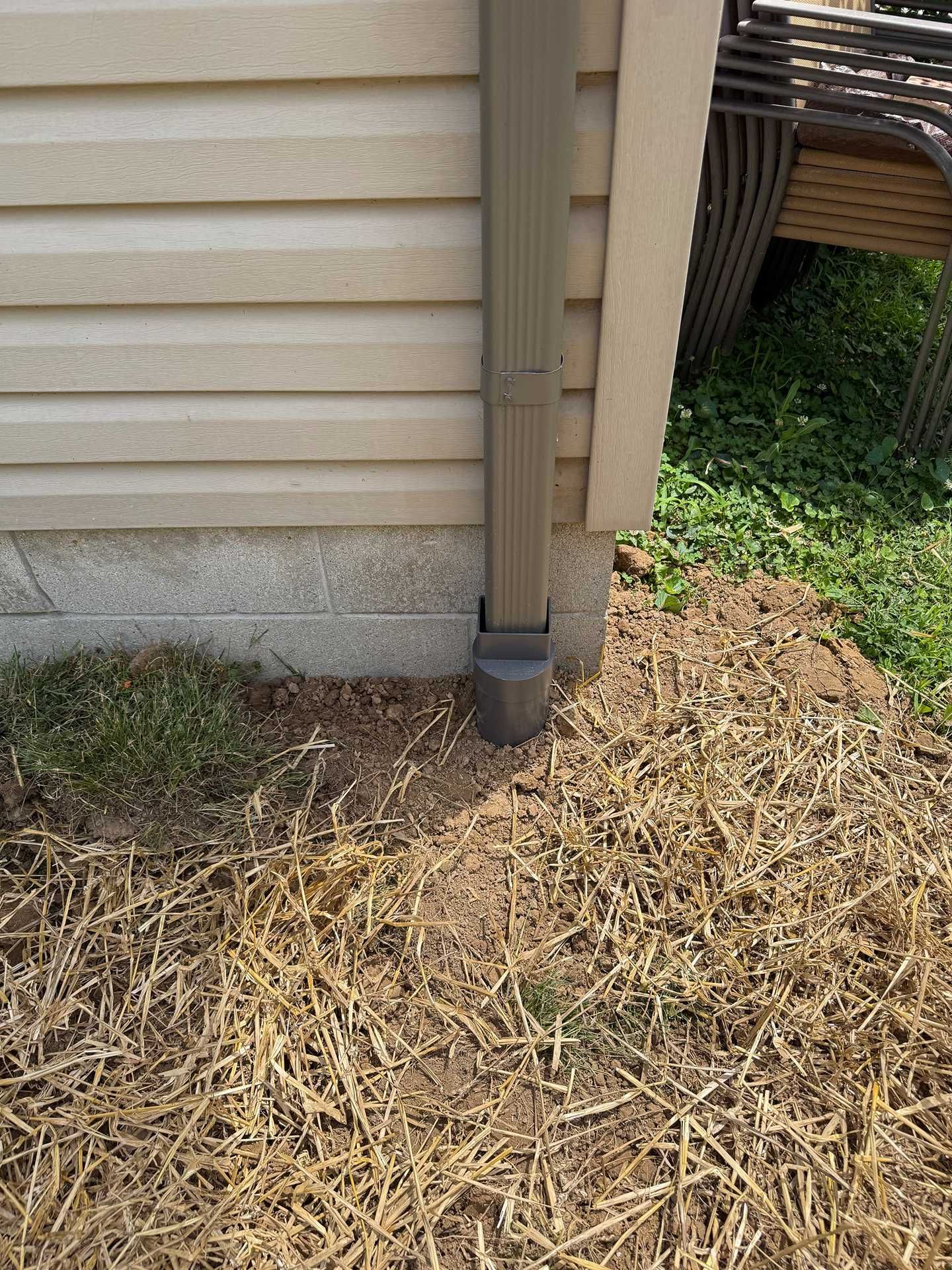 House corner with beige siding, downspout, and dry grass/mulch along the foundation.