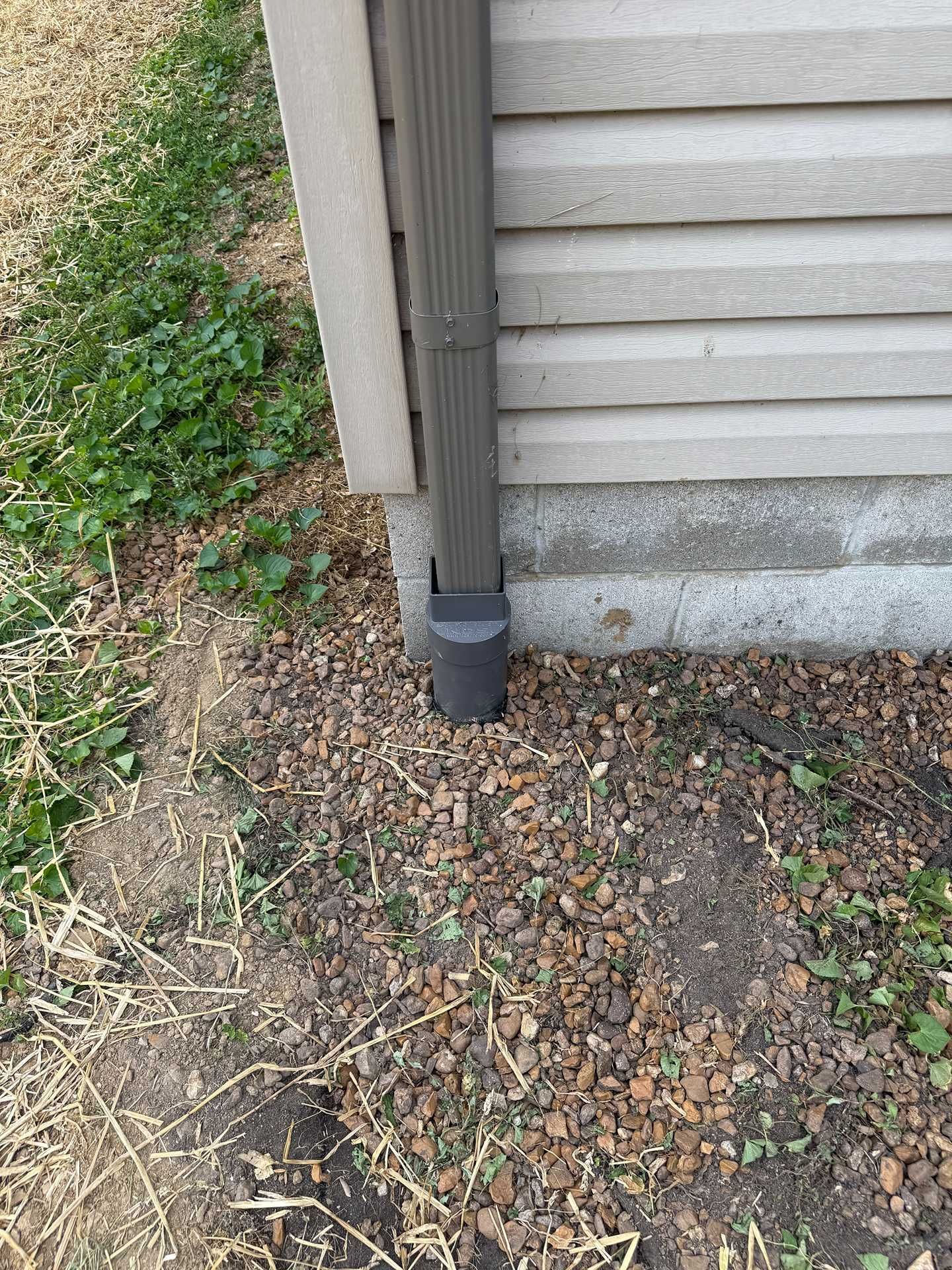 House downspout and siding beside a gravel foundation edge with weeds and dirt path