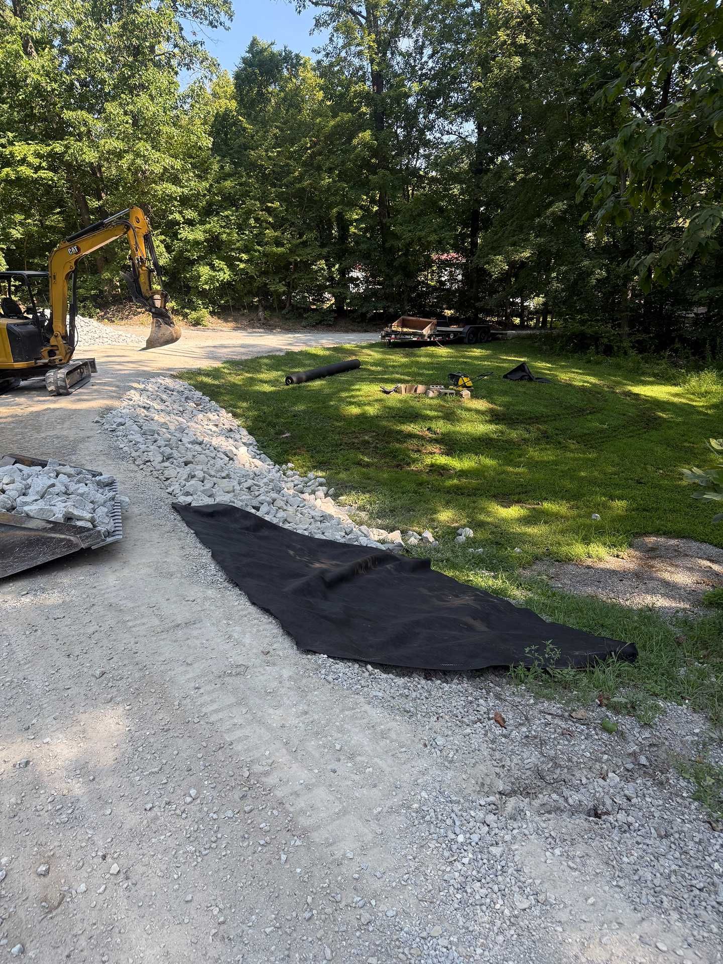 Gravel driveway under construction with an excavator, black tarp, and trees beside a grassy yard
