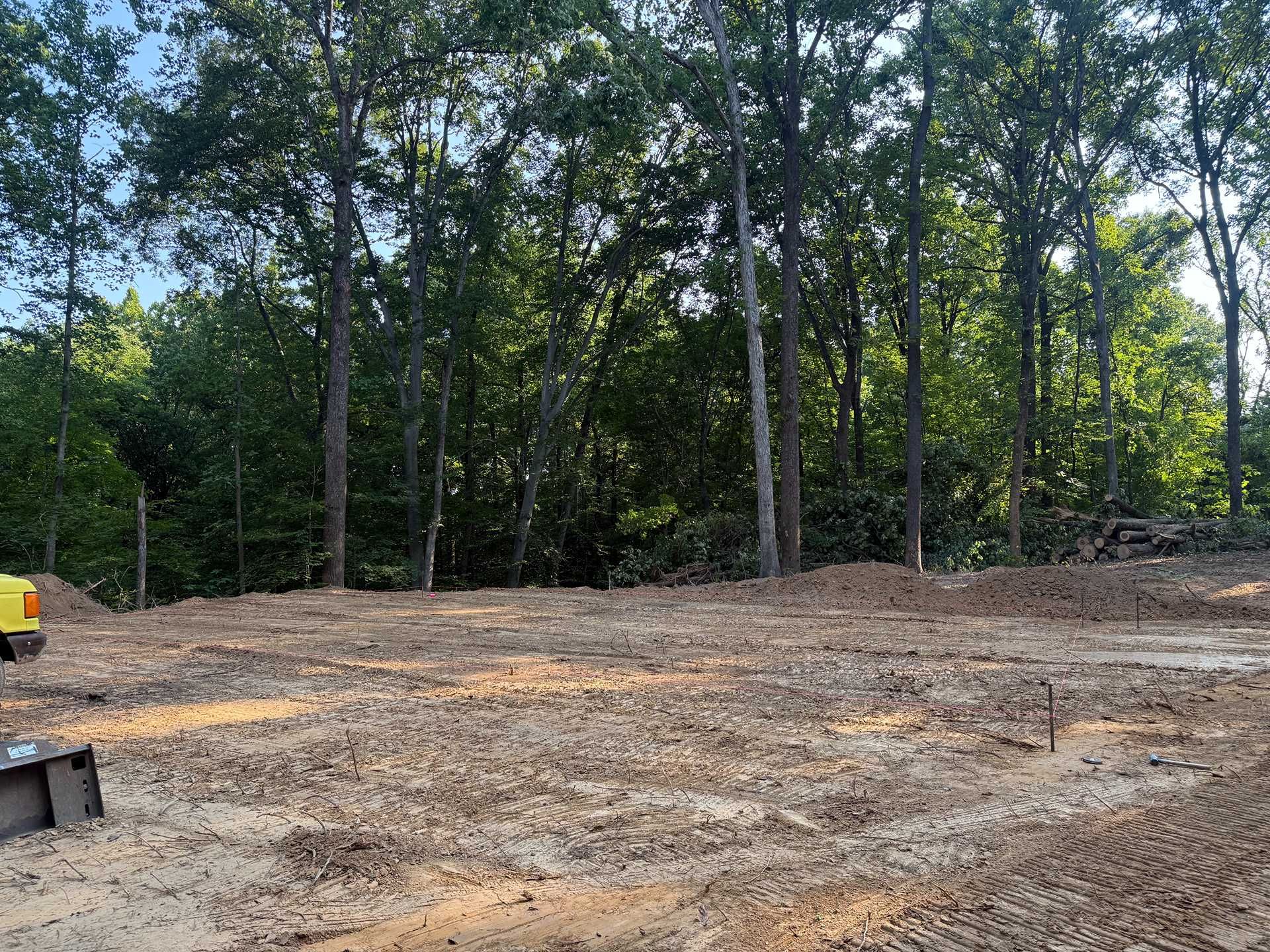 Cleared dirt lot bordered by tall trees, with a partial yellow excavator at left.