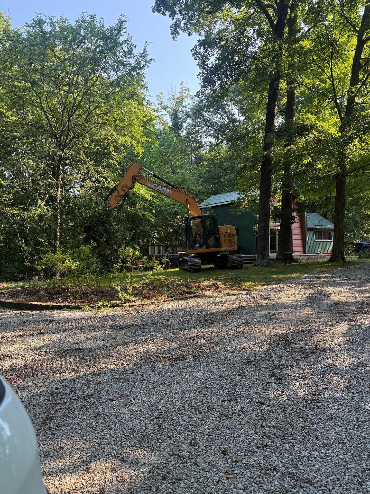 Yellow excavator in a wooded yard beside a small red building on a gravel driveway.