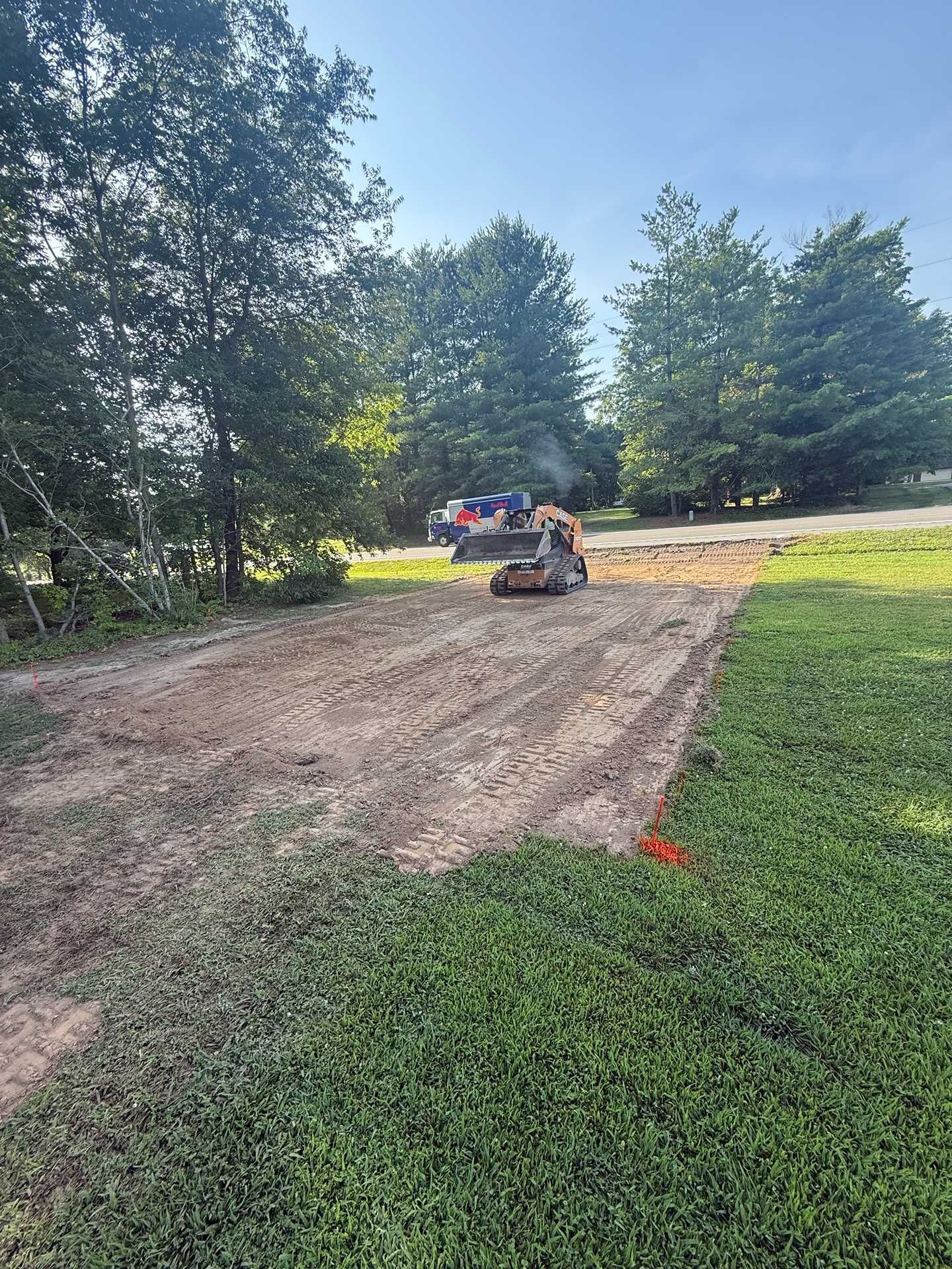 Backhoe leveling a dirt path in a grassy yard, with trees and a truck in the background.