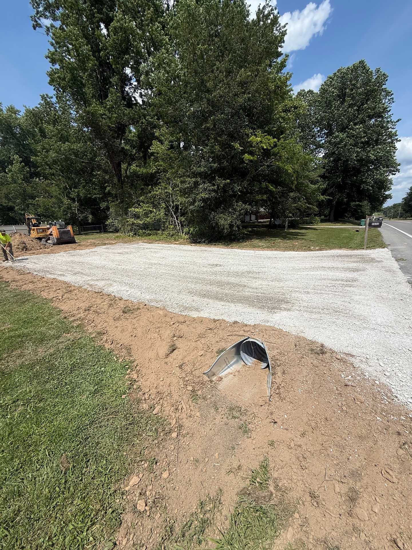 Gravel driveway beside trees, with a small drainage pipe in a dirt ditch in the foreground.