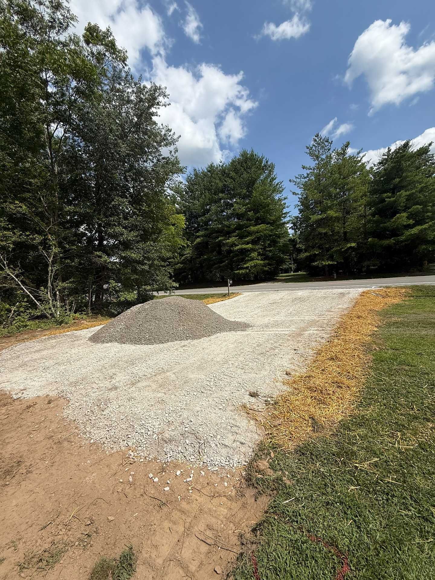 Gravel driveway under construction beside trees, with a pile of gravel and blue sky overhead