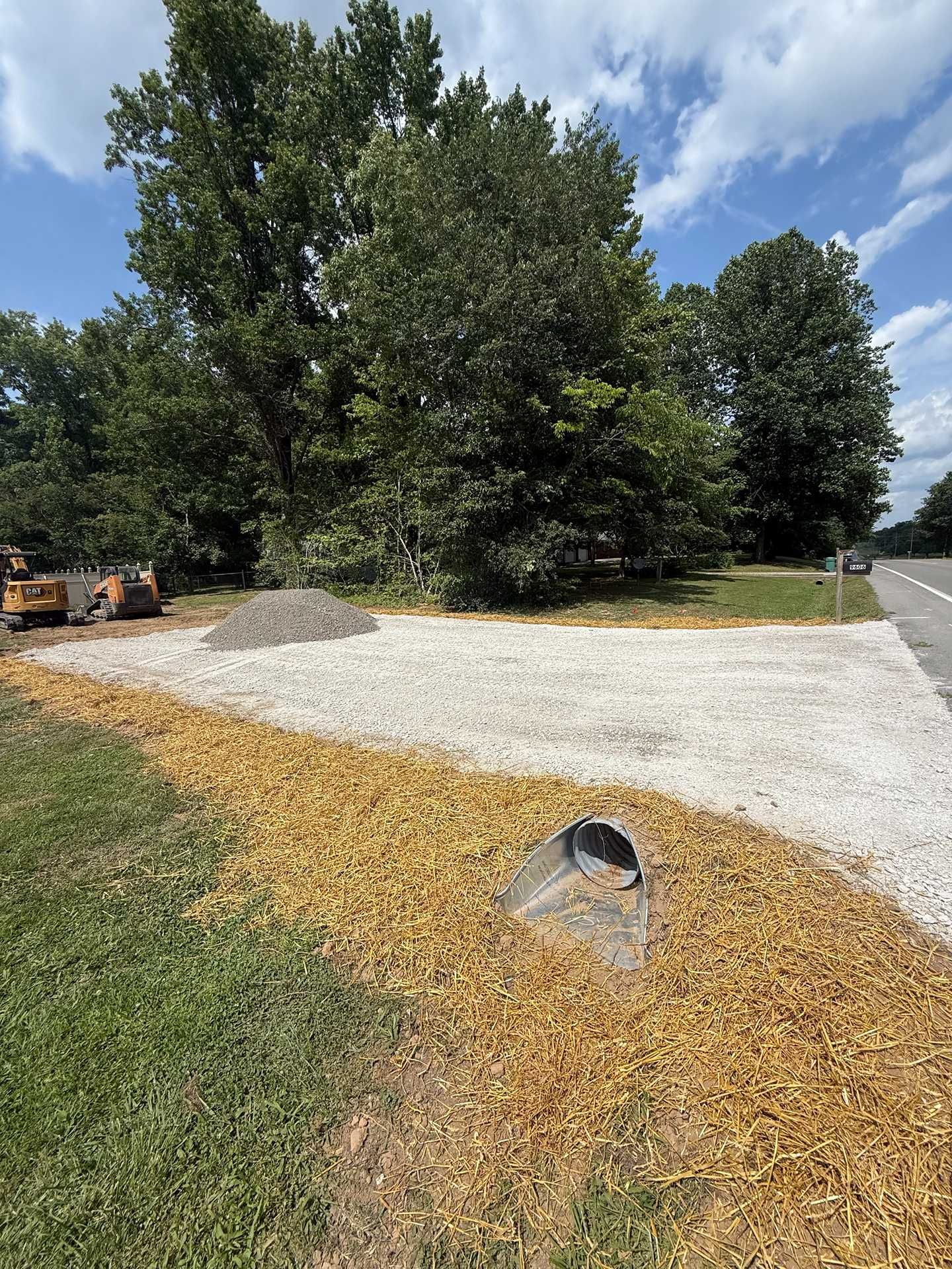 Gravel driveway with a culvert drain, bordered by grass and large trees under a blue sky