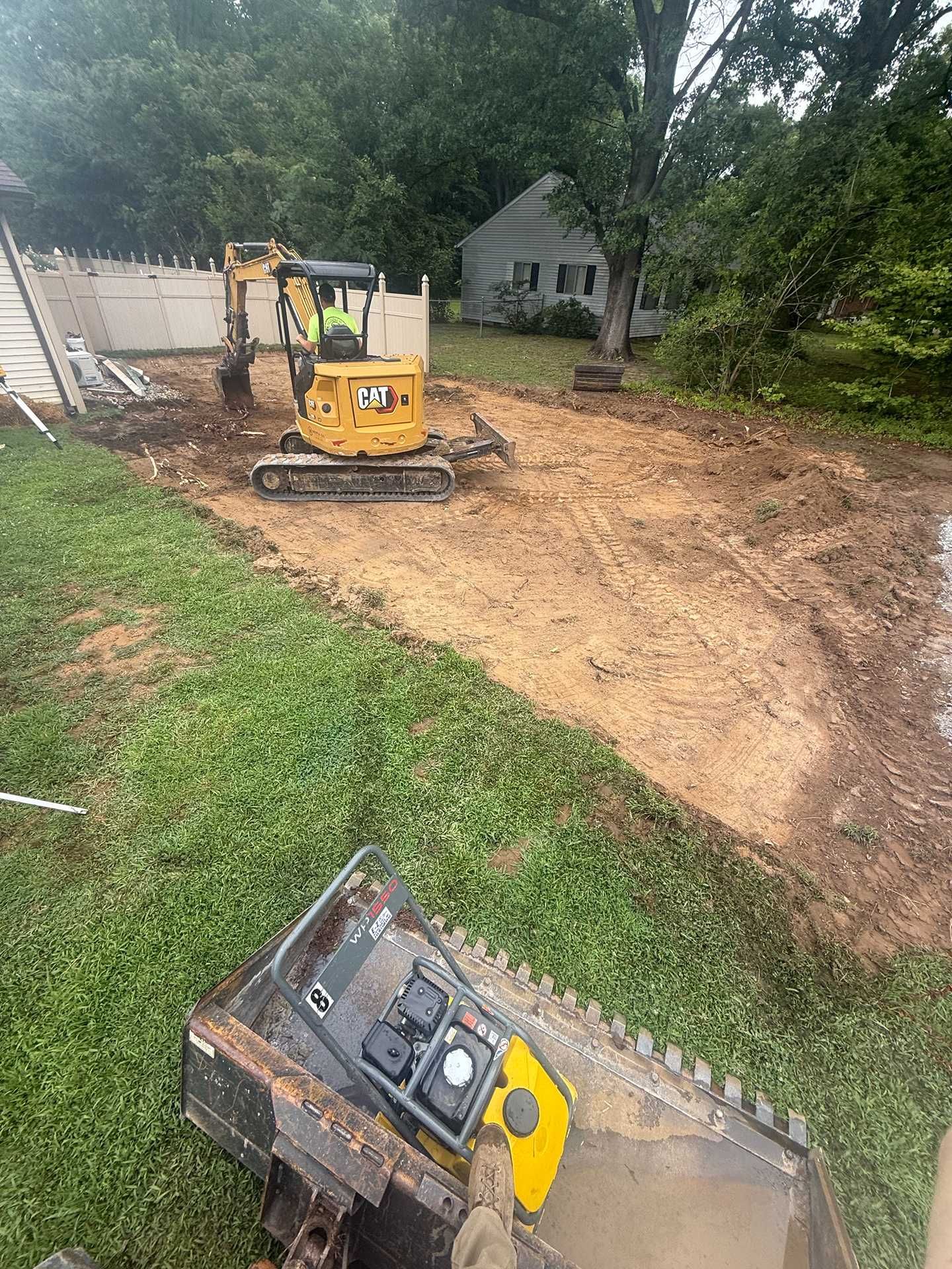 Yellow mini excavator on a muddy construction site near a house and trees.
