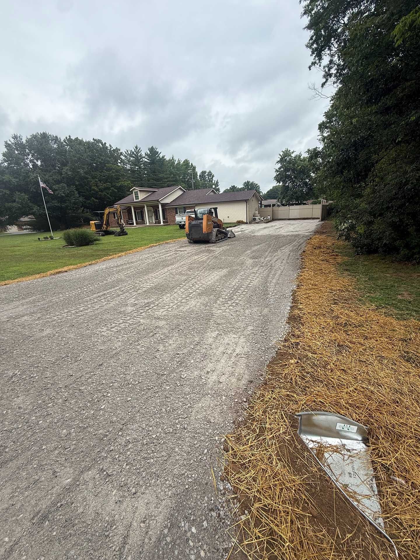 Gravel driveway with a small excavator near a house and garage on a cloudy day
