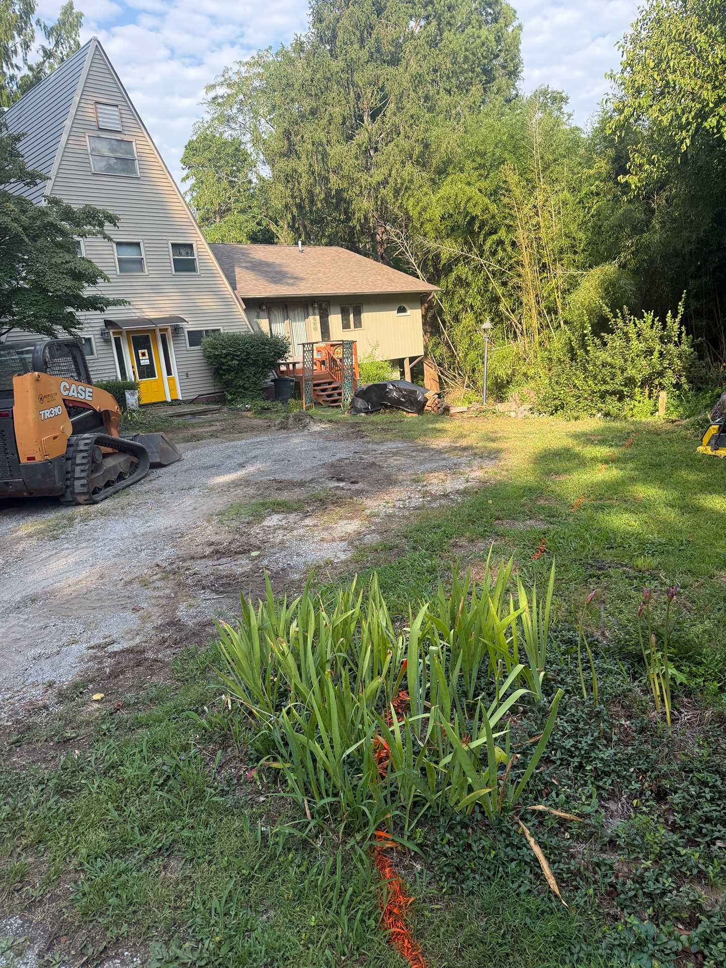 Cabin with gravel driveway, parked construction vehicle, and lush garden in a wooded yard