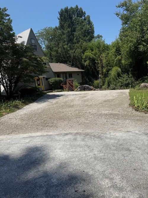 Gravel driveway leading to a house surrounded by tall green trees on a sunny day