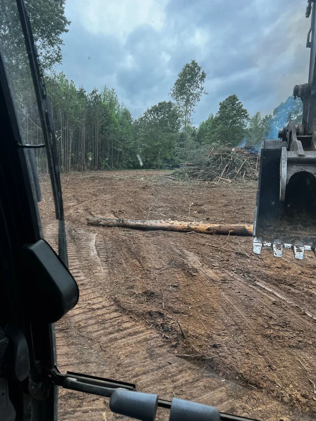 An excavator clearing a wooded area. A felled log lies in the center of the brown dirt. Trees and a pile of debris are visible.