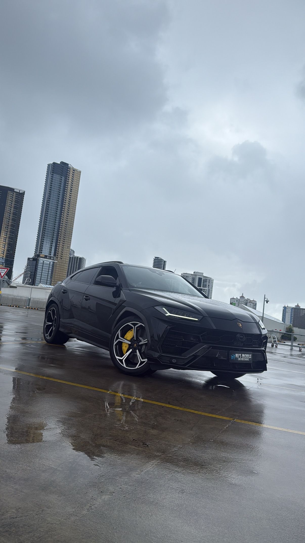 Black Lamborghini Suv Parked on Wet Asphalt in Front of Tall Buildings — MJ's Mobile Car Mechanic In Austinmer, NSW