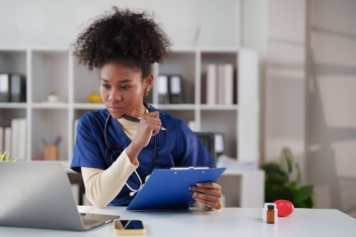 Female nurse looking at laptop while writing on clipboard at MDCredentialing