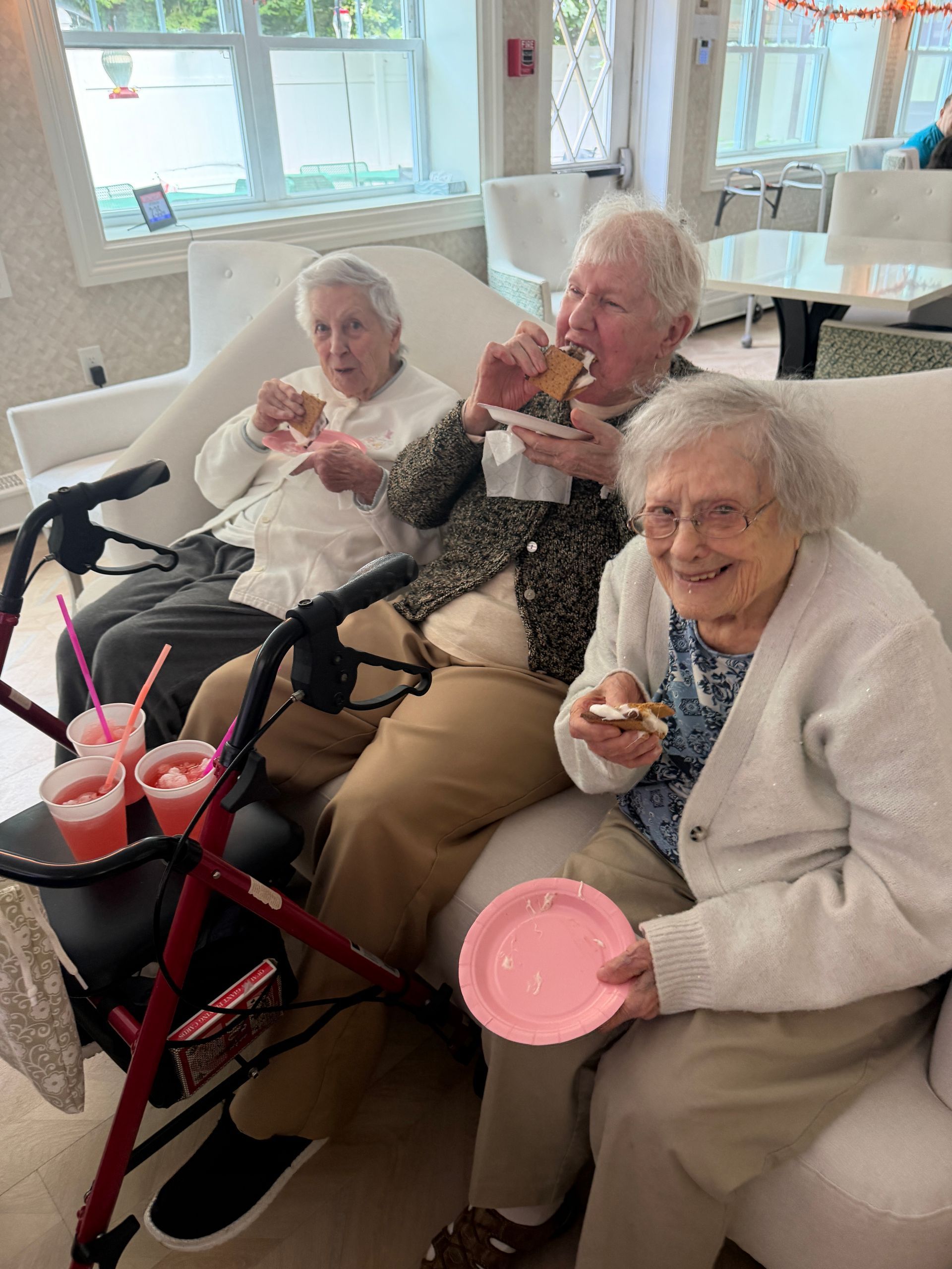 Three elderly people sit in a bright room, smiling and eating dessert with walkers beside them.