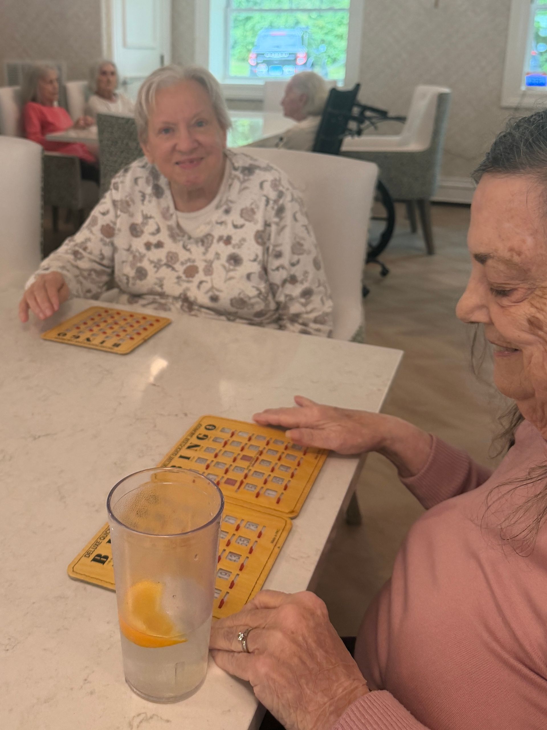 Two people playing bingo at a table in a bright room, with bingo cards and a drink on the table.