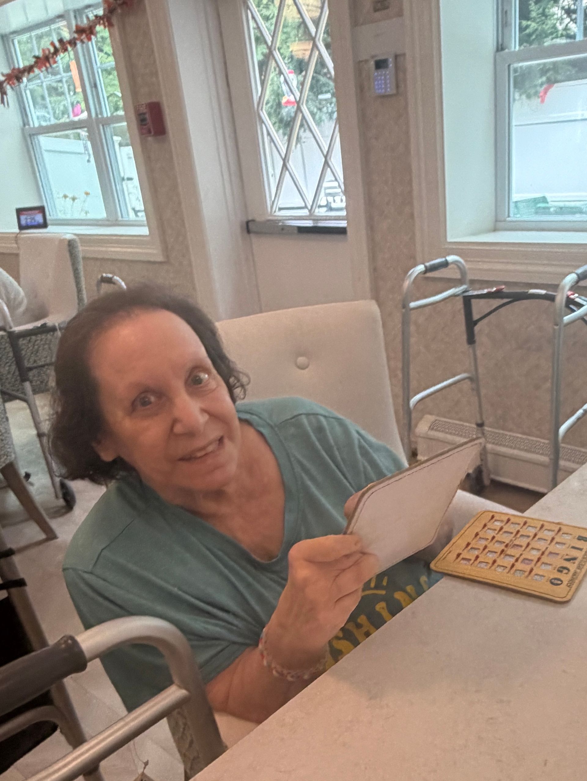 Woman seated at a table, holding a card and smiling in a bright room with windows and a walker nearby