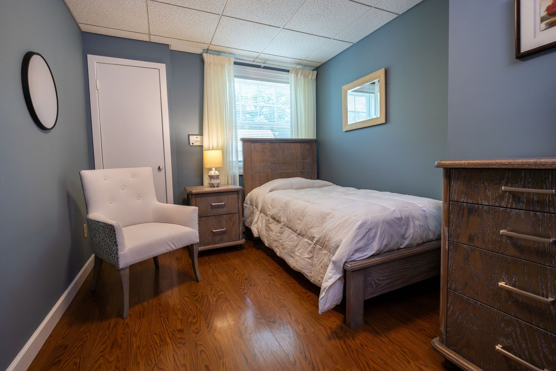 Small bedroom with twin bed, white chair, blue walls, wood floor, and dresser by the window.