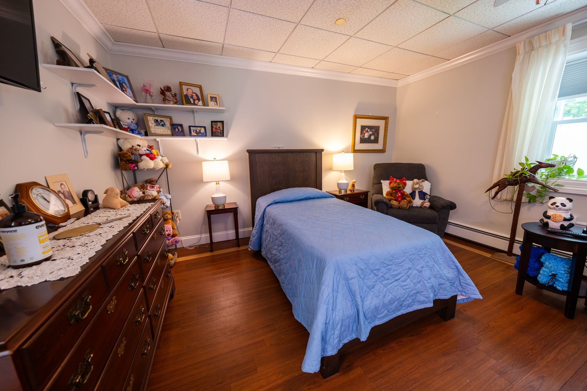 Cozy bedroom with blue bedspread, wooden dresser, shelves, lamp, and sunlight through a window