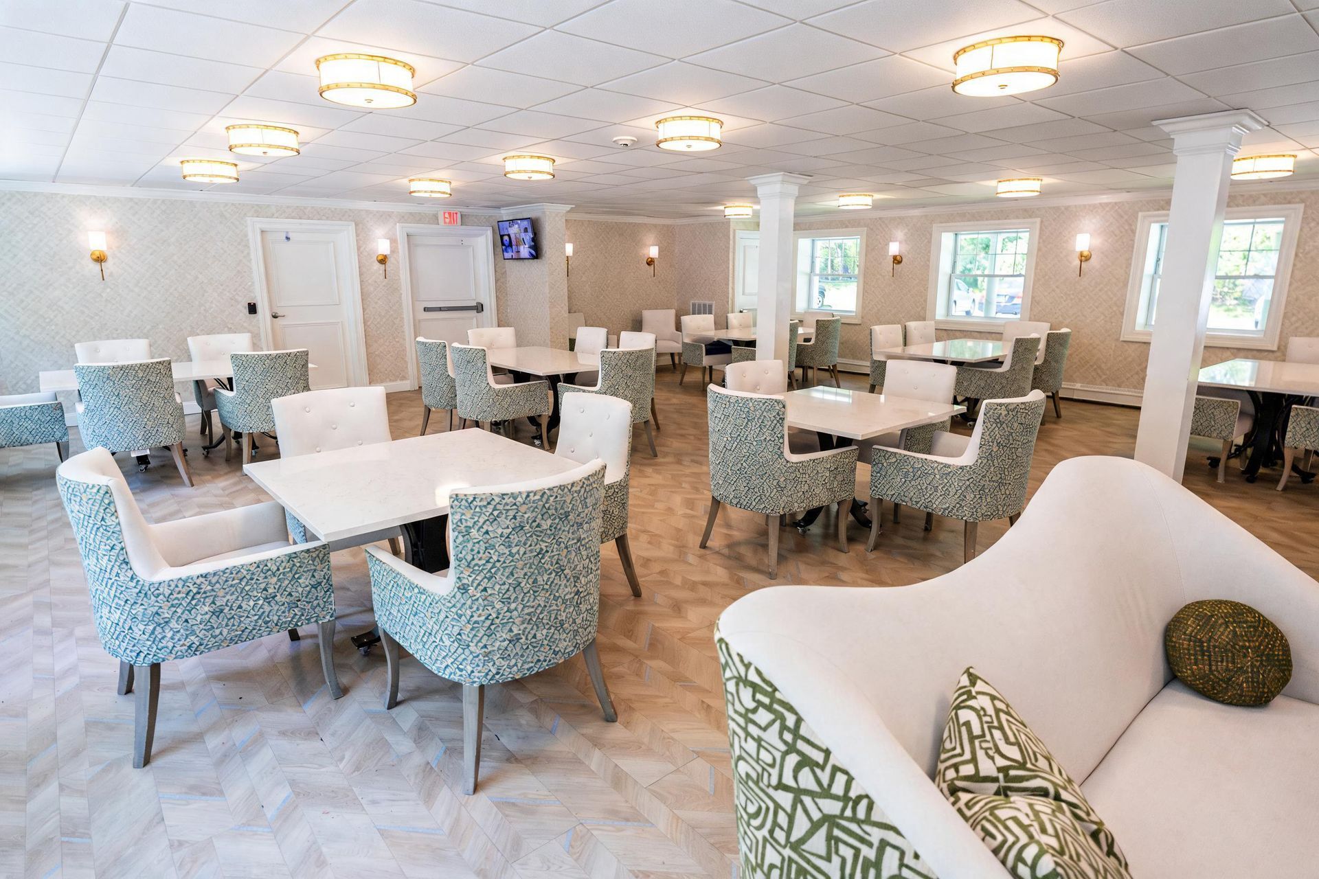 Bright, modern dining room with white tables, pale blue chairs, and patterned sofas under ceiling lights.