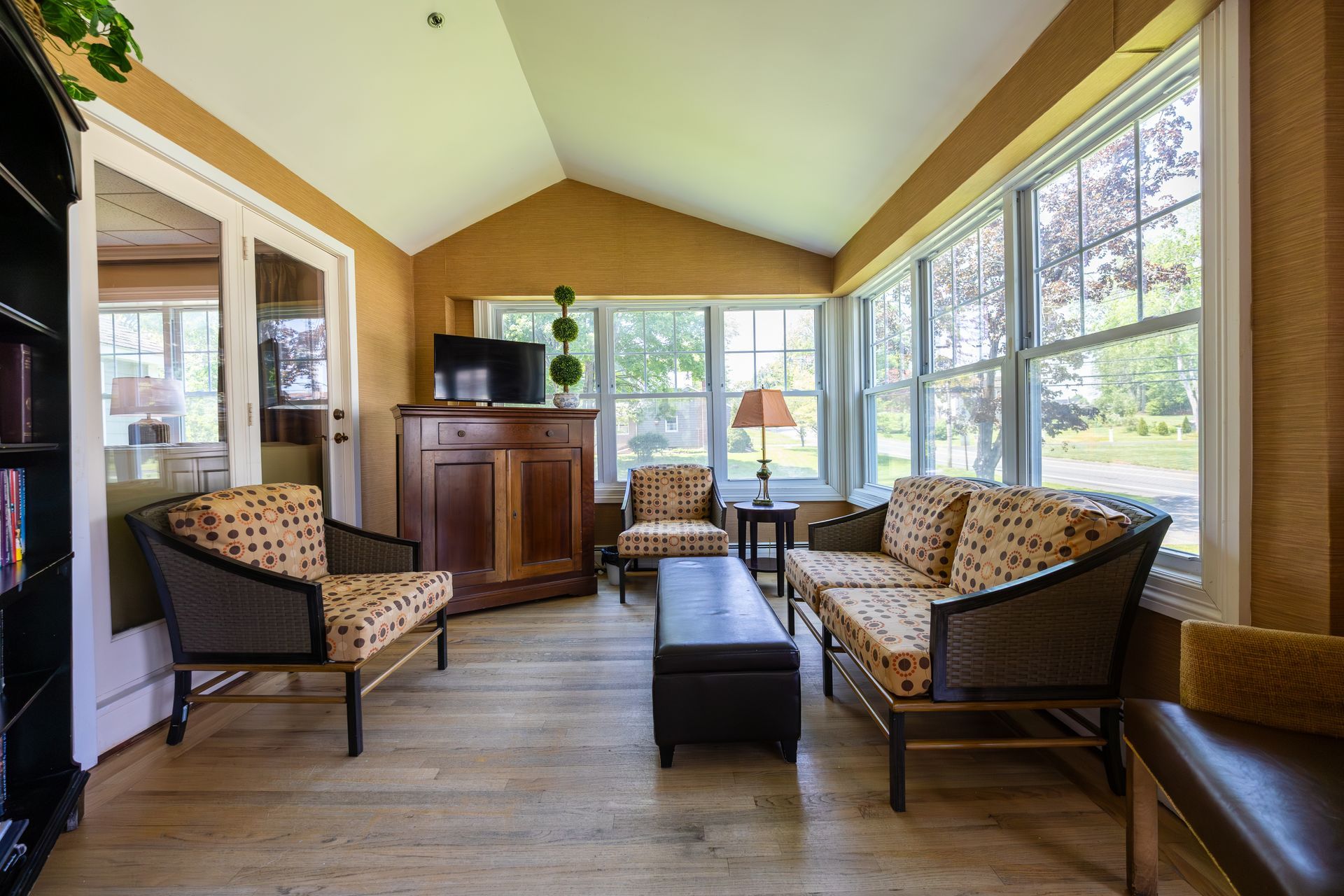 Sunroom lounge with wicker chairs, a dark ottoman, wood cabinet, and large windows overlooking greenery