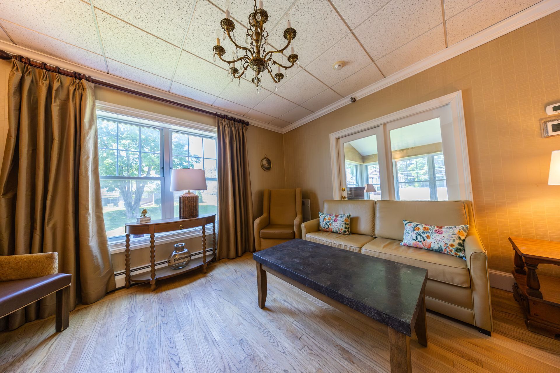 Warm living room with beige sofa, coffee table, curtains, and large windows with daylight.