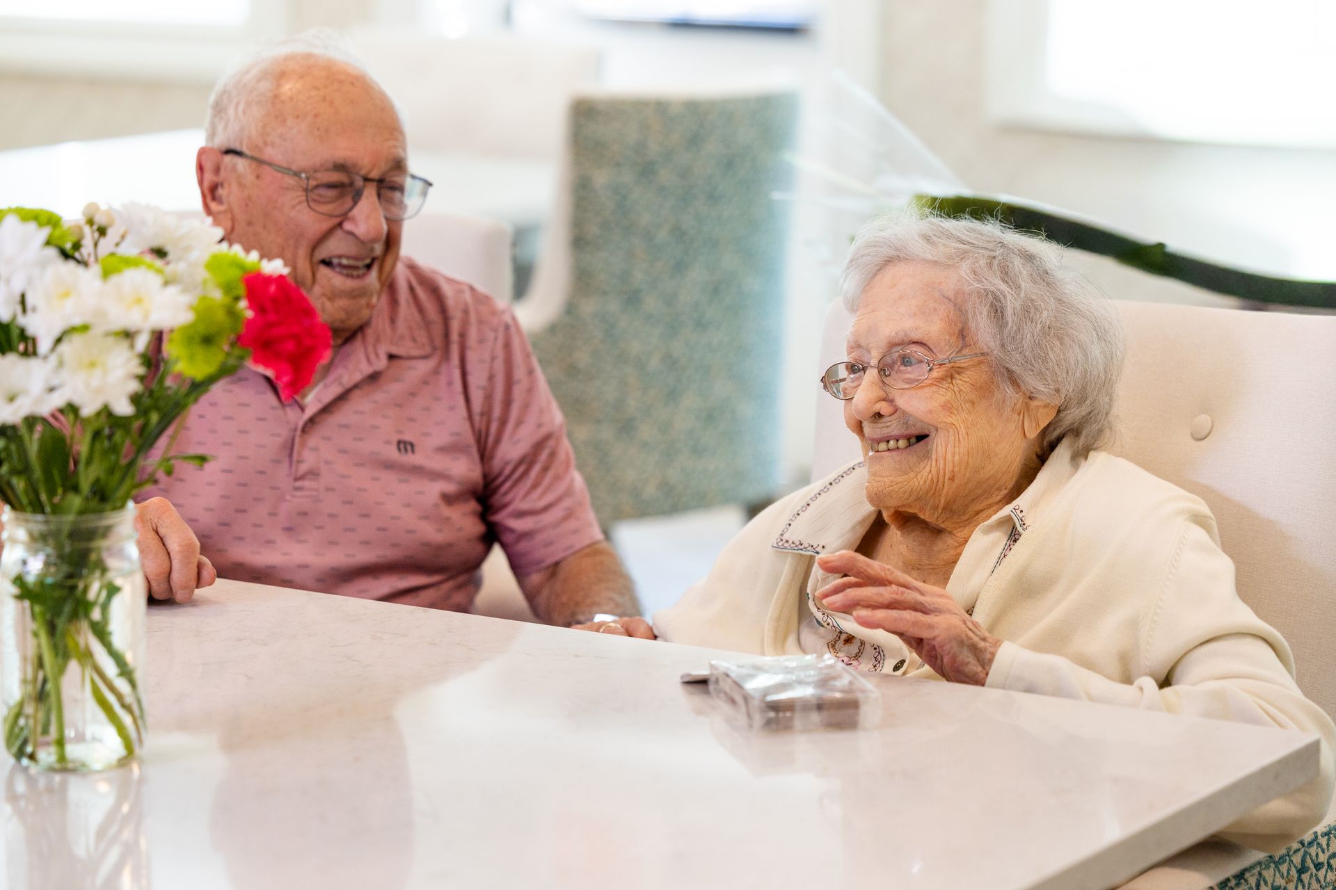 Two elderly people smiling and talking at a bright dining table with flowers in a vase.