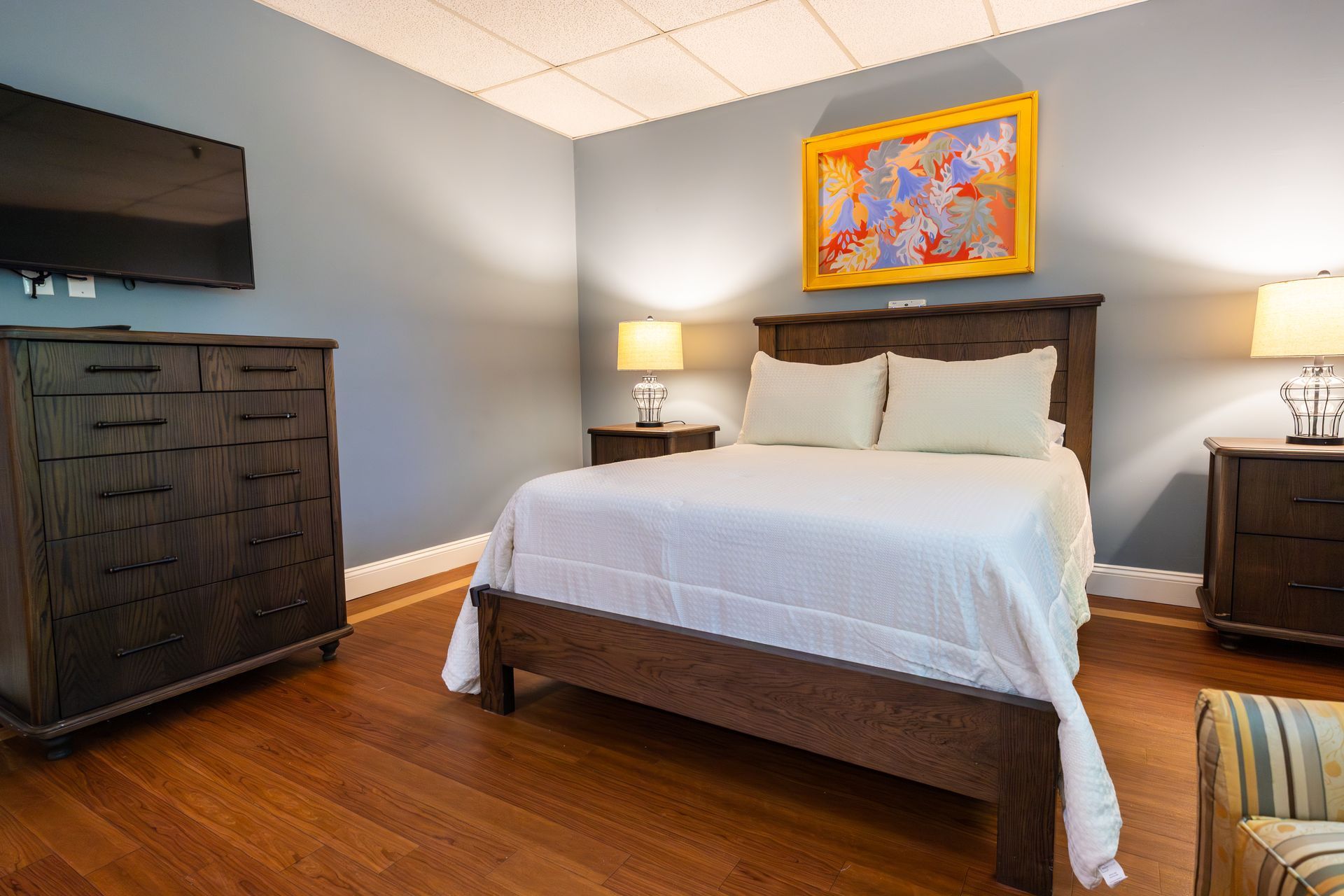 Cozy bedroom with a made bed, hardwood floors, bedside lamps, and colorful wall art.