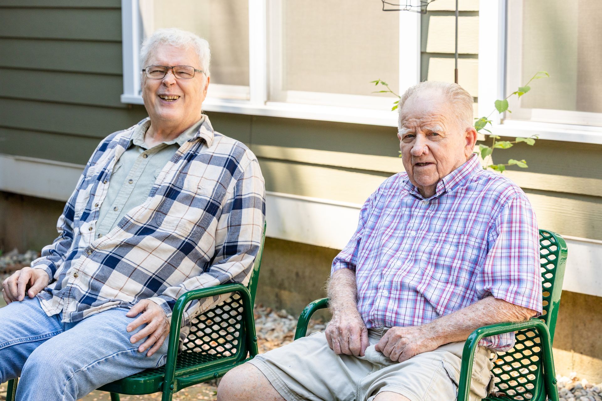 Two elderly men sit in green chairs outside a house, one in plaid shirt smiling and the other in a striped shirt.