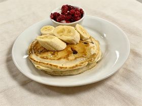 Pancakes topped with banana slices and served with a bowl of raspberries on a white plate.