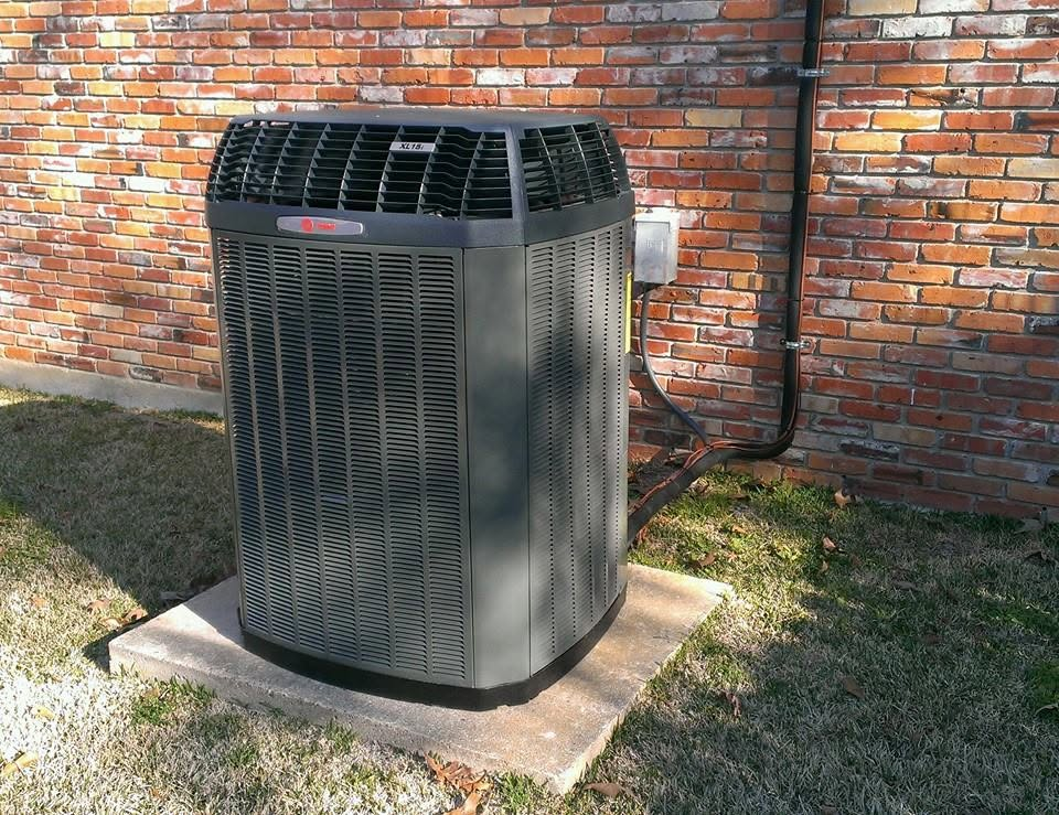 Outdoor HVAC unit beside a brick wall on a concrete pad, surrounded by grass.