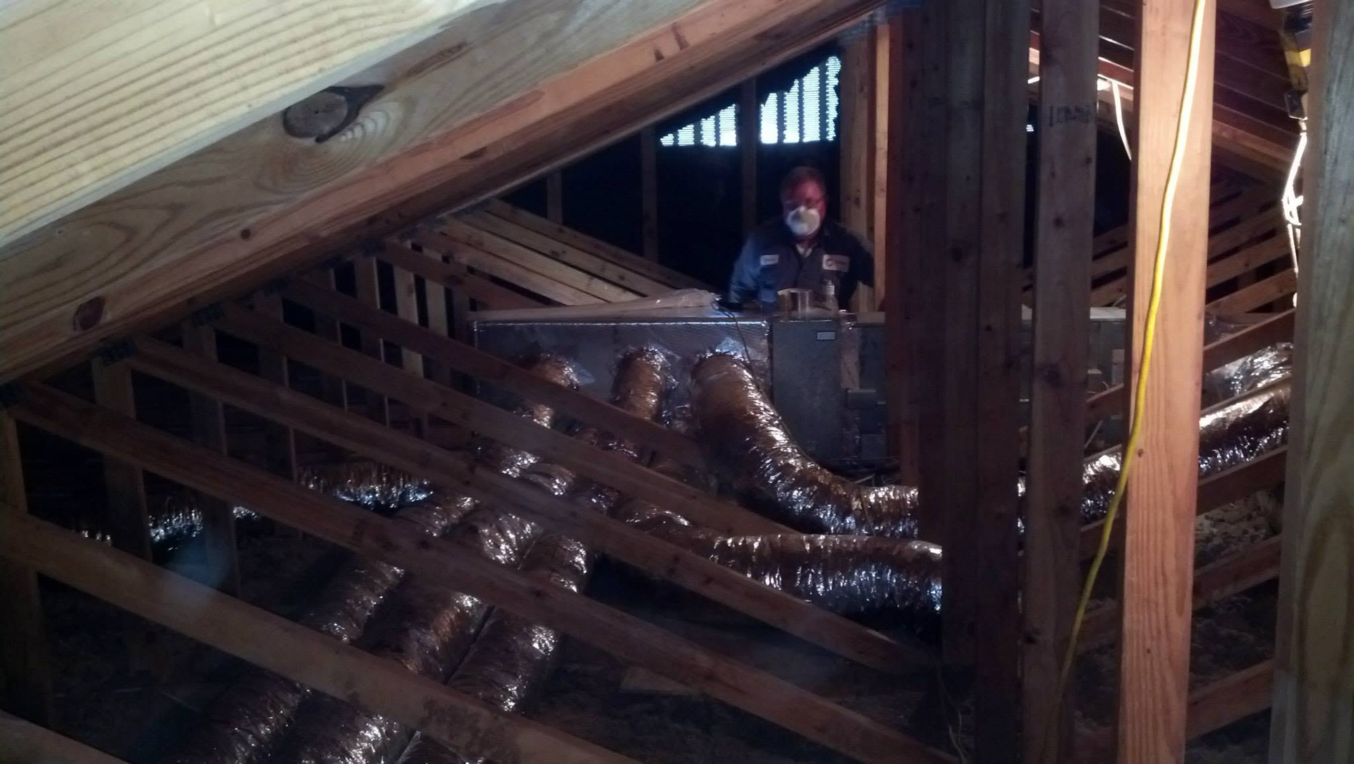 Person crouching in an attic beside shiny insulated ductwork and exposed wooden beams.