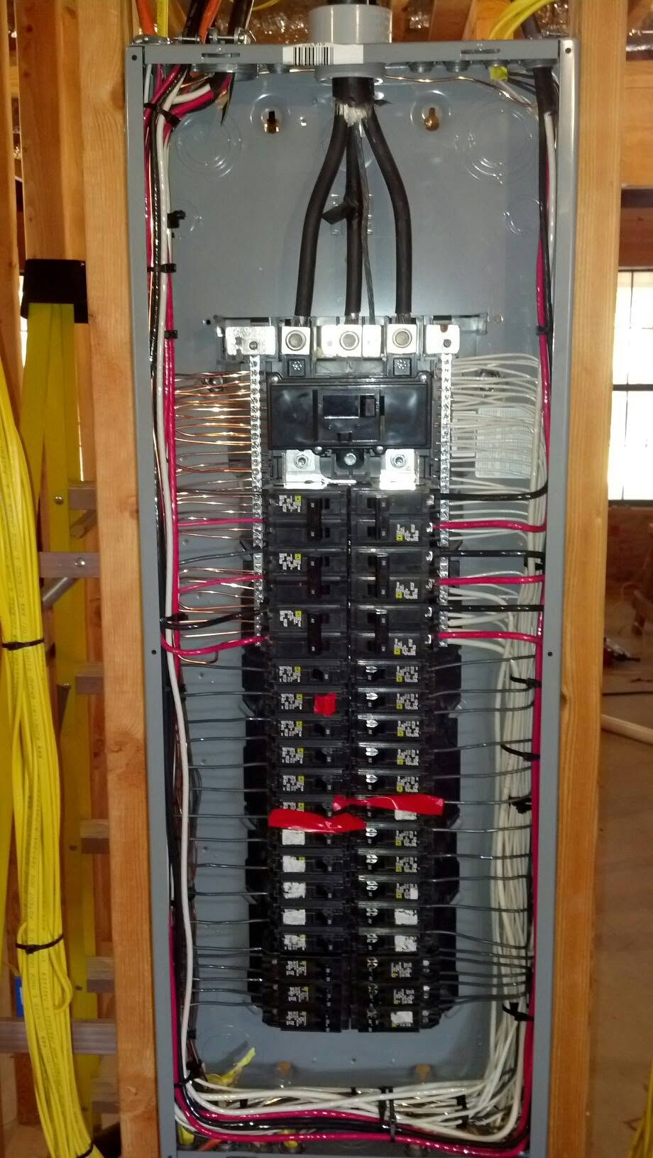 Open electrical breaker panel with rows of circuit breakers and red and black wires inside a wooden wall cavity