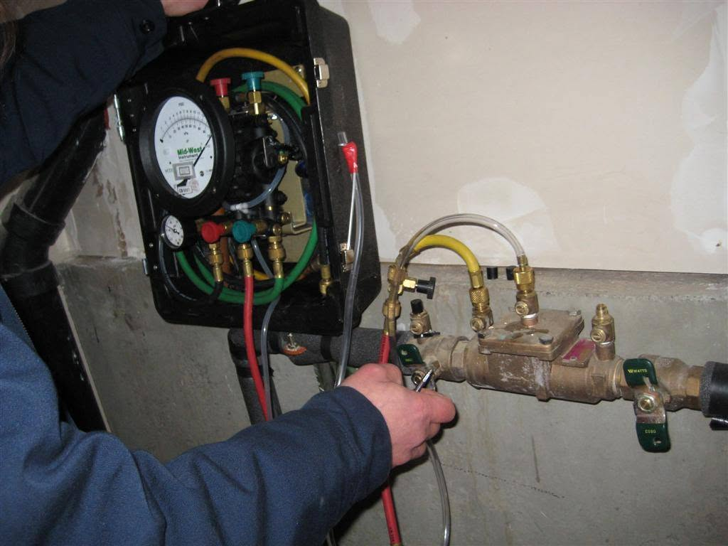 Technician checking a wall-mounted plumbing valve assembly with gauges and colored hoses.