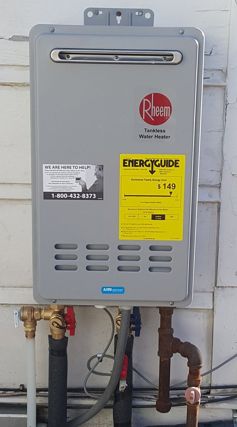 Wall-mounted gray tankless water heater with yellow energy label and exposed pipes on a white wall